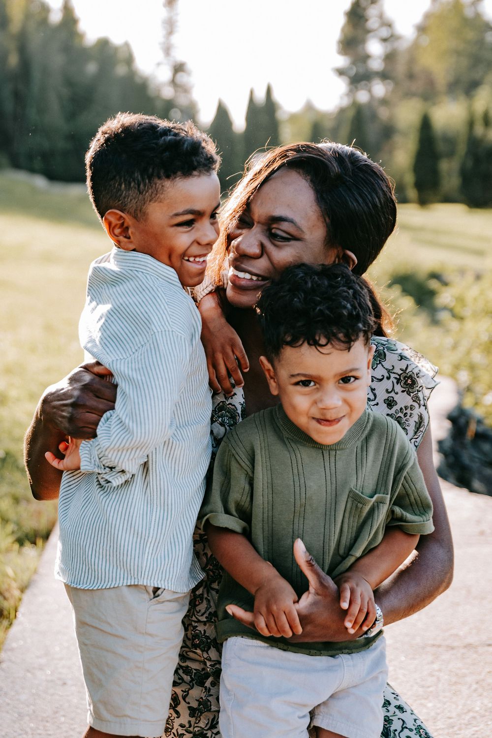 A joyful mother hugs her two young sons outdoors, all smiling warmly in a sunlit green park setting.