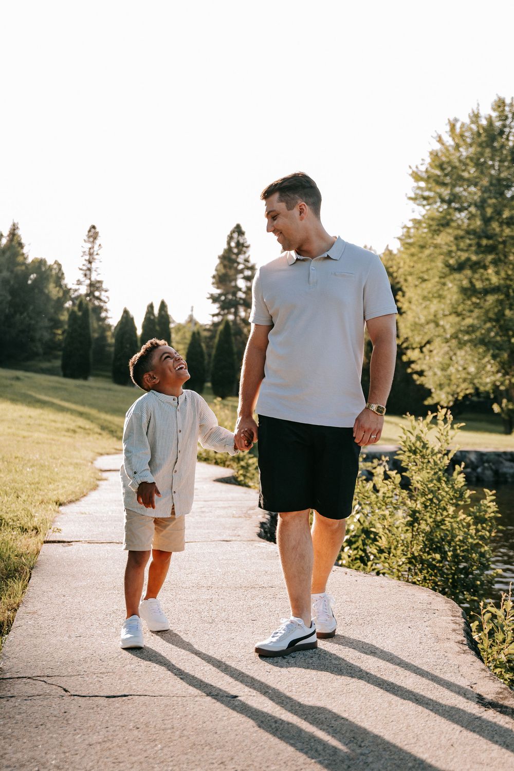 A man and young boy walk hand in hand on a sunny park path, smiling at each other in casual summer outfits.