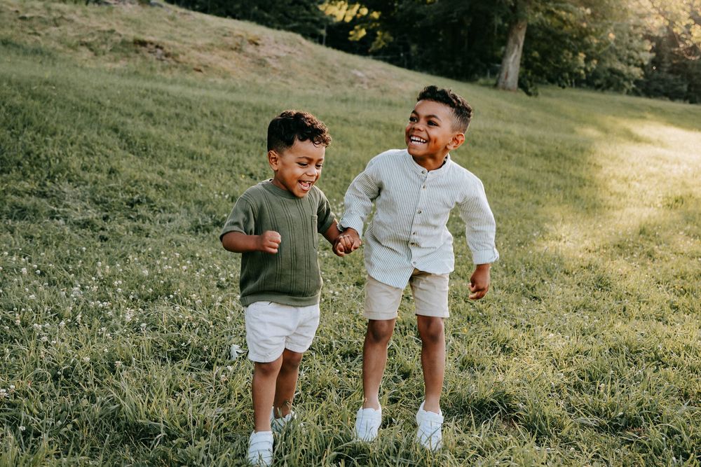 Two joyful young boys running and holding hands on a green grassy hill in warm sunlight.