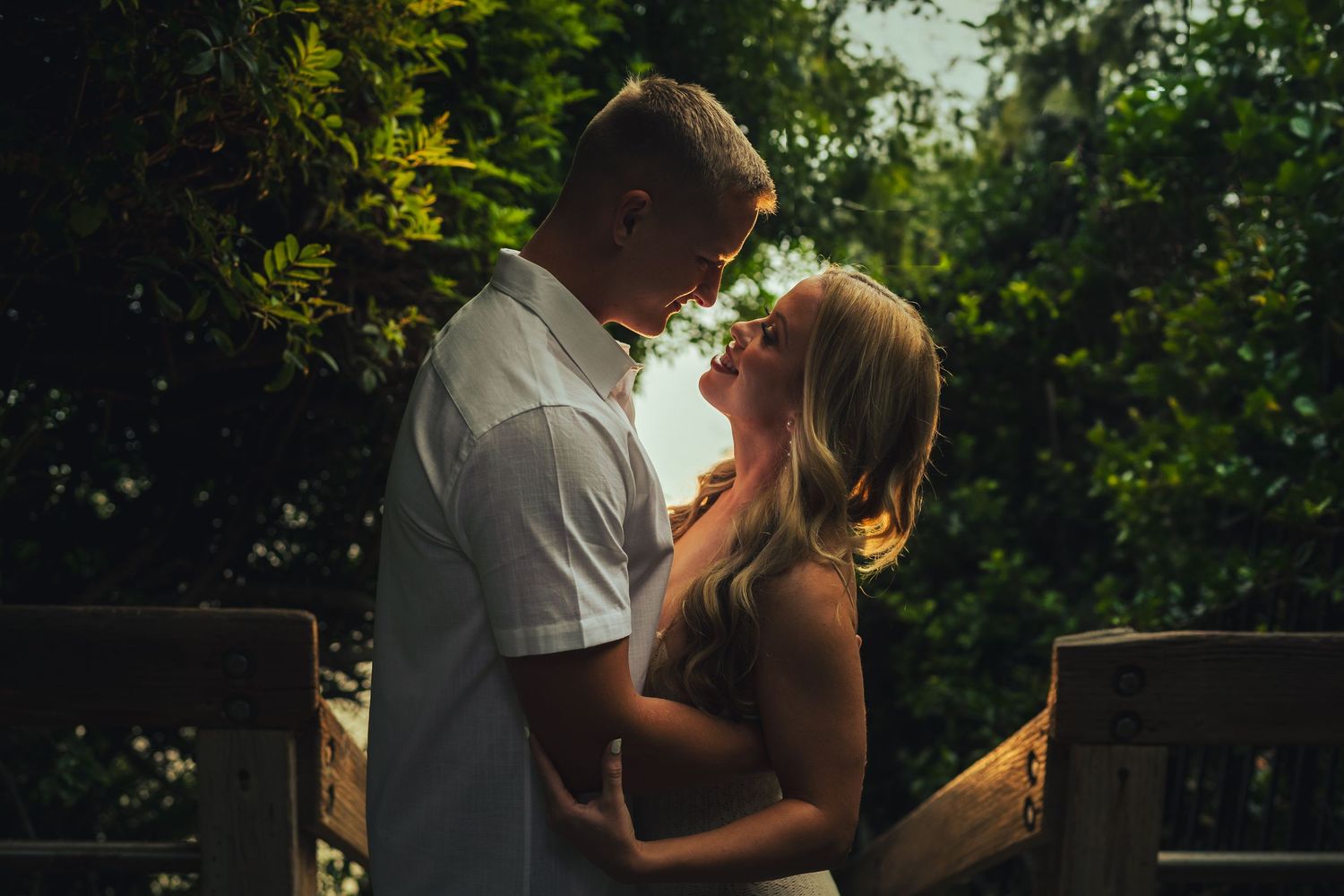 Romantic couple embraces in warm backlit sunset glow with lush greenery in background.