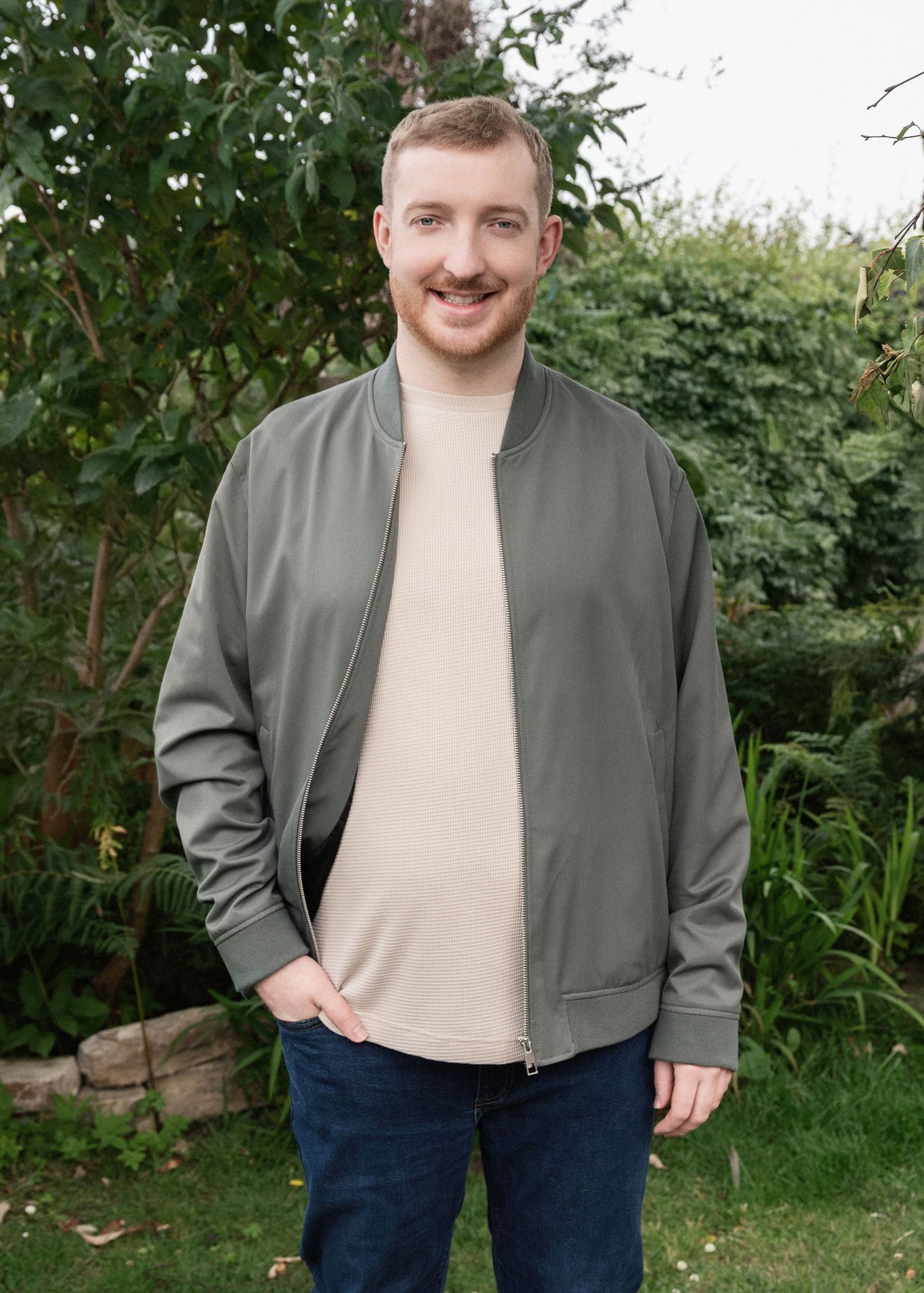 Someone wearing a casual olive bomber jacket and beige t-shirt stands smiling in a garden setting with green foliage.