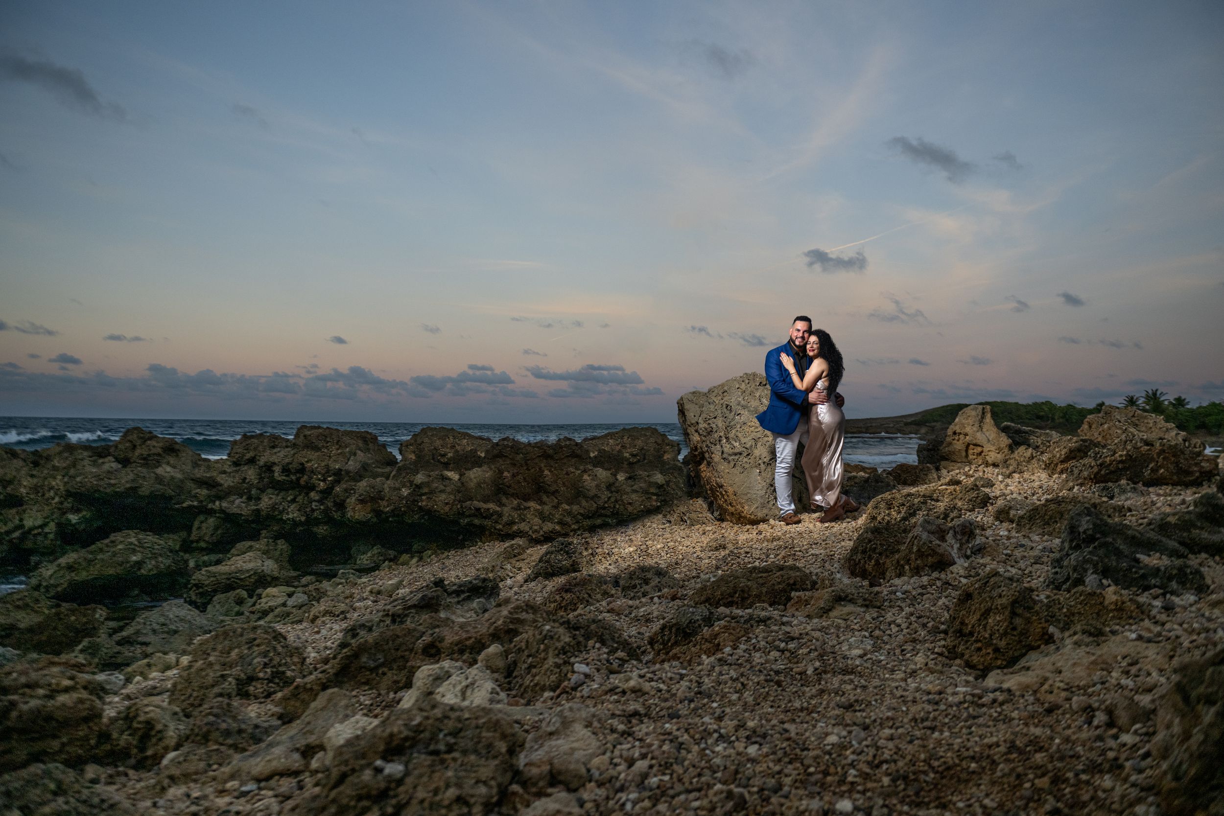 Couple sits atop rocky coastal terrain at sunset during their engagement photoshoot.