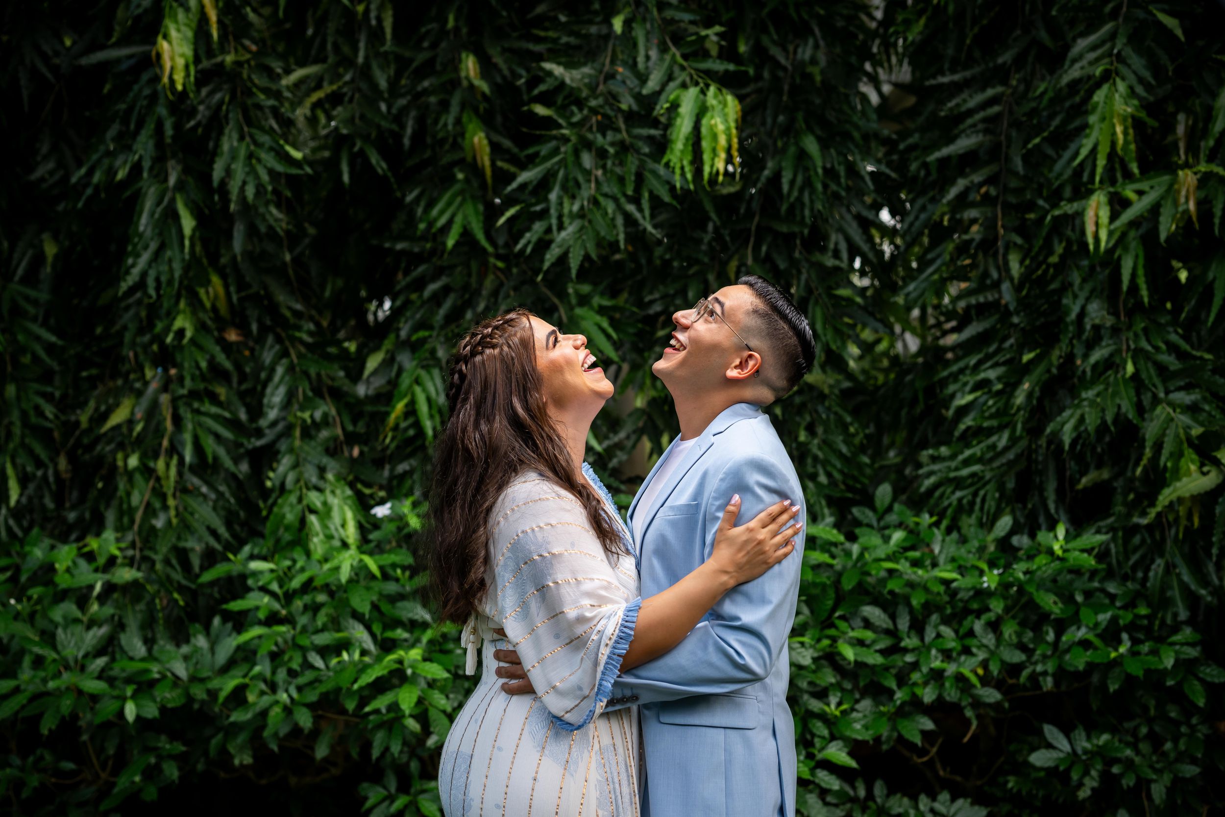 Two people in matching white and blue outfits embrace beneath lush green foliage at dusk for romantic couples photos.