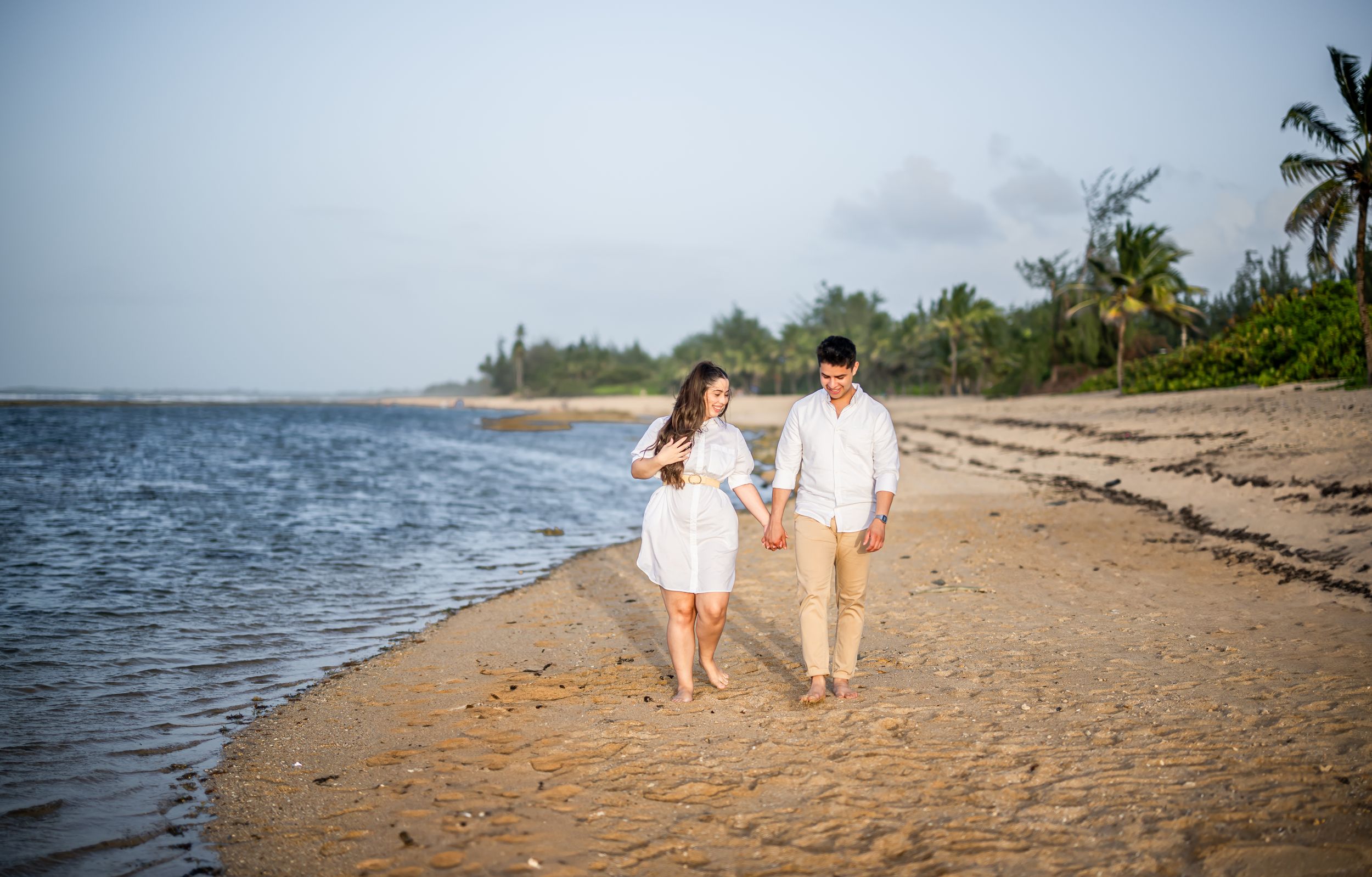 Couple walking hand in hand along a scenic tropical beach with palm trees and ocean waves at sunset.