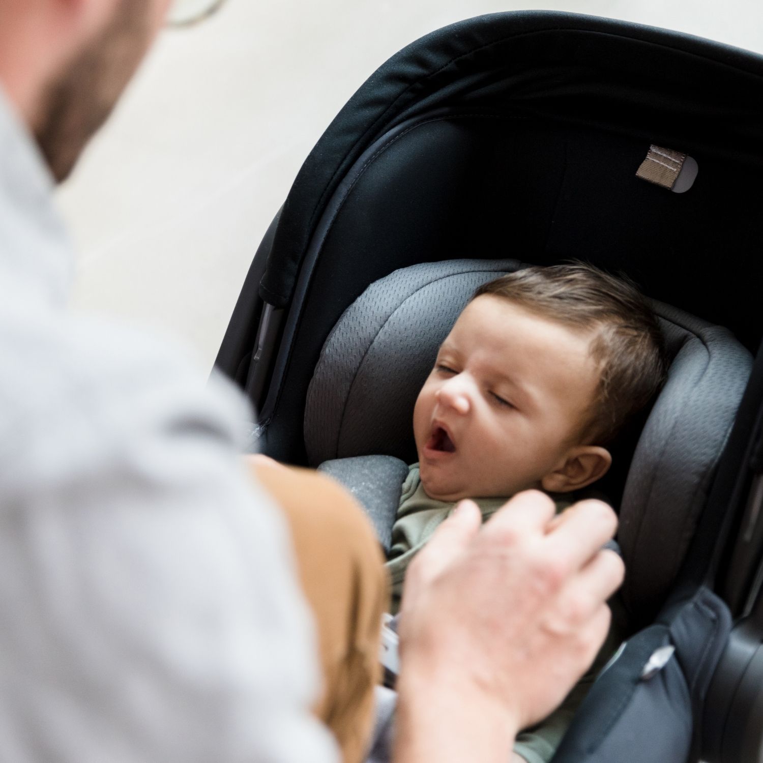 Newborn baby yawning in a carseat. A parent is next to him.