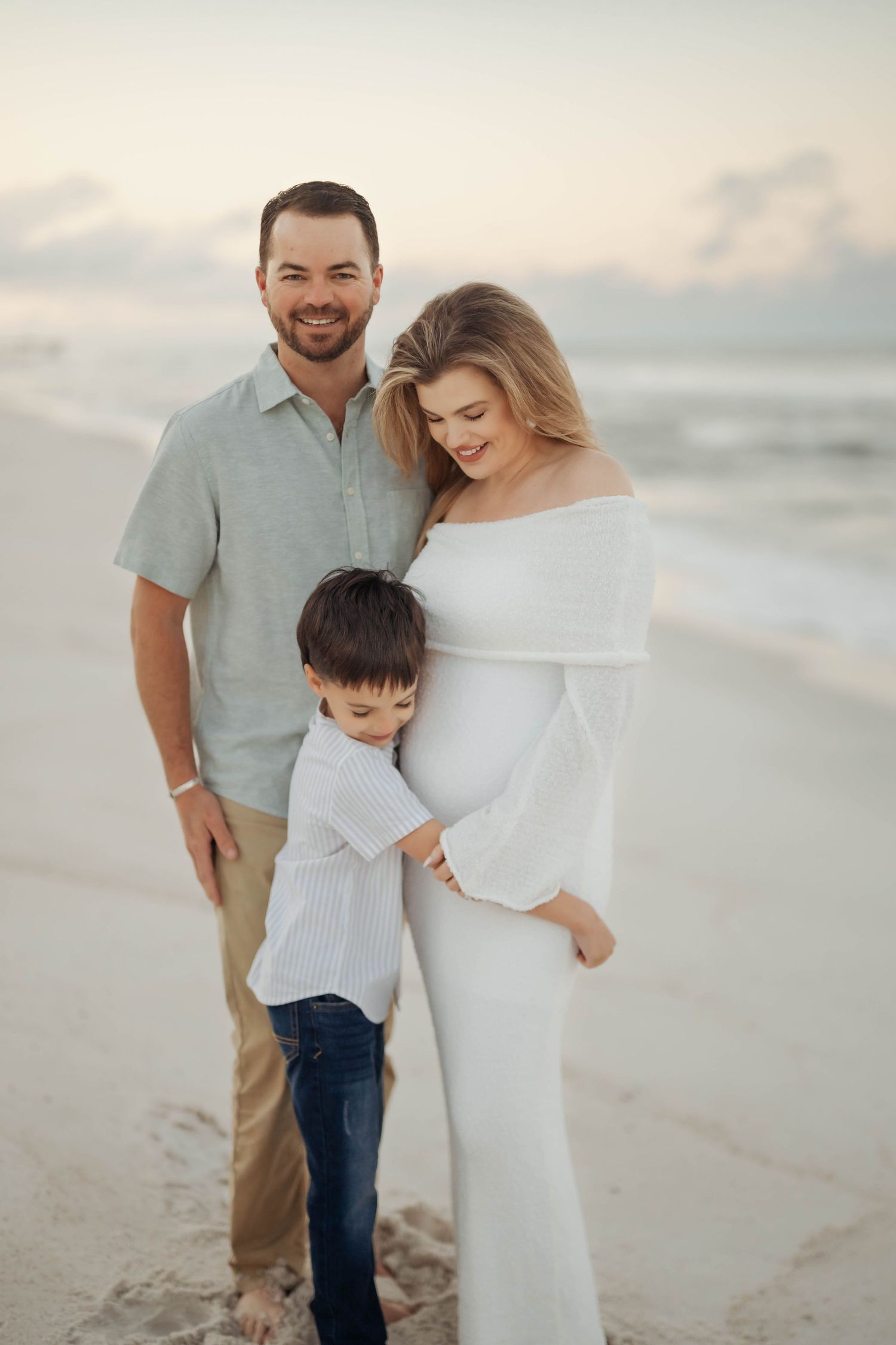 A family in coordinating white and neutral outfits poses together on a beach at sunset enjoying a casual photo session.