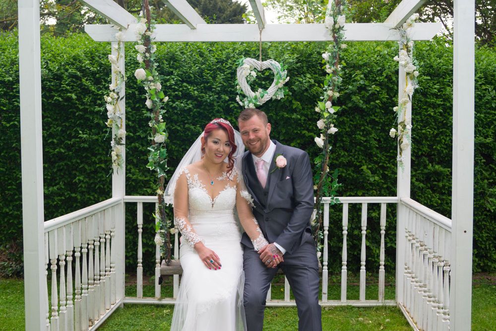 A wedding couple poses under a white garden arbor decorated with flowers and a heart.