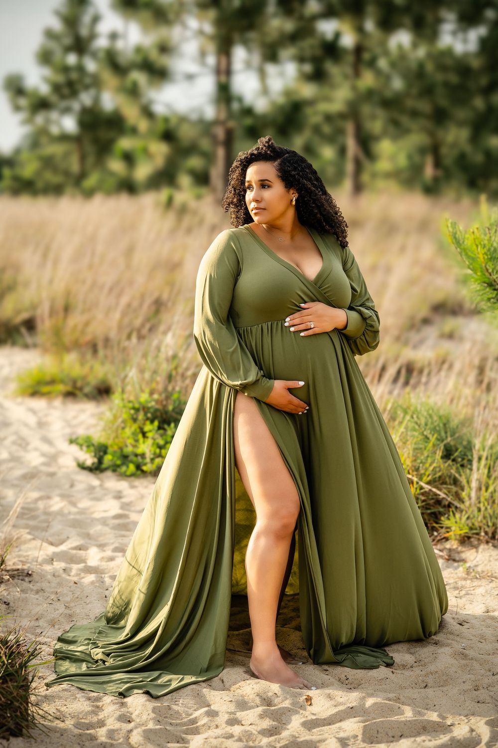 A flowing olive green dress with long sleeves worn in a natural outdoor setting with tall grass and pine trees.