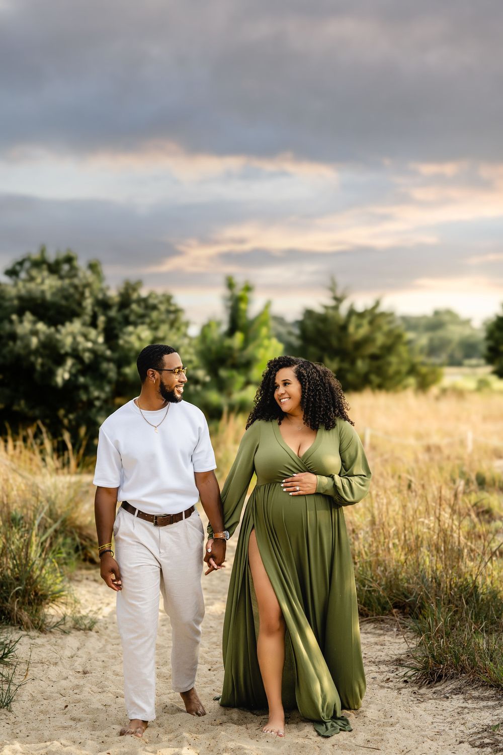 Couple walks hand-in-hand along a sandy path through tall grass at sunset with dramatic cloudy skies above.