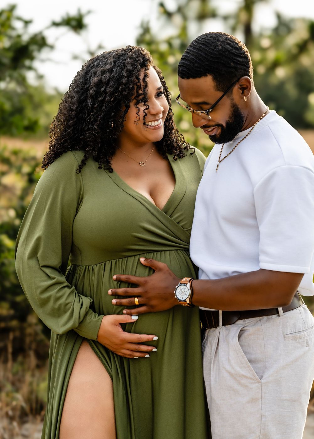 Expecting couple in olive green dress and white shirt embrace lovingly during maternity photo session outdoors at sunset.