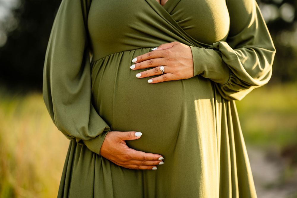 Close-up of pregnant belly in flowing olive green maternity dress with hands gently cradling the bump outdoors.