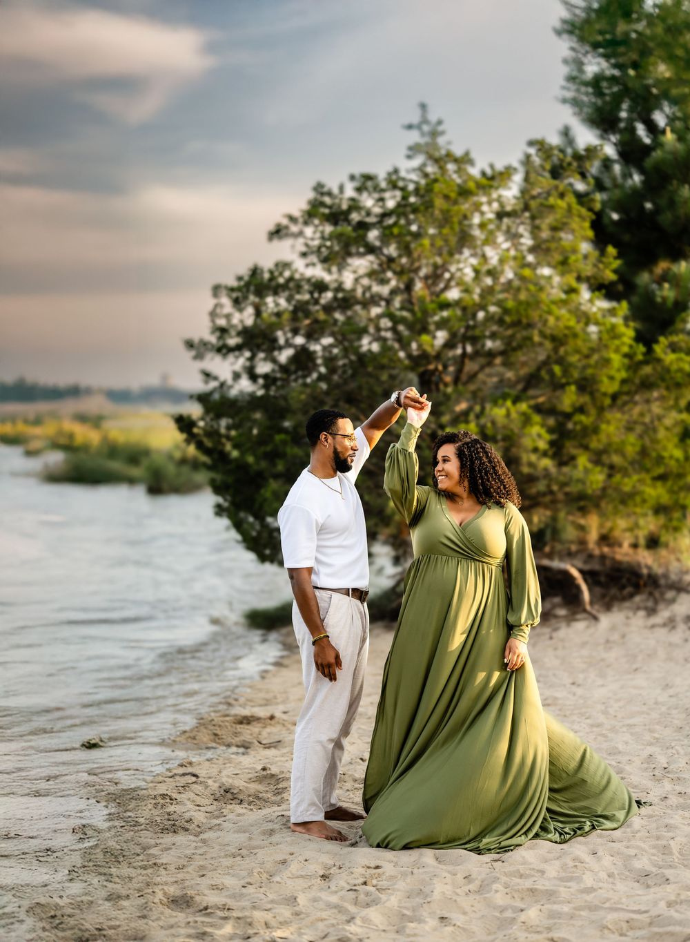 A romantic beach photoshoot featuring a flowing olive green dress and white outfit against a scenic waterfront at sunset.