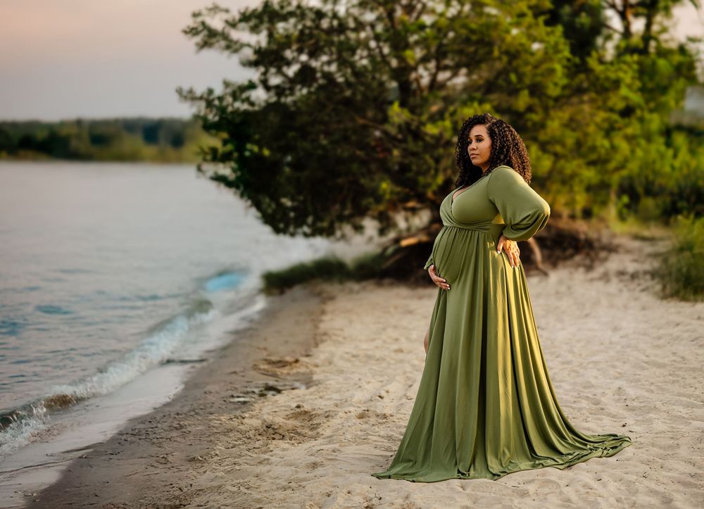 A flowing sage green maternity gown on a beach at sunset with water and trees in the background.
