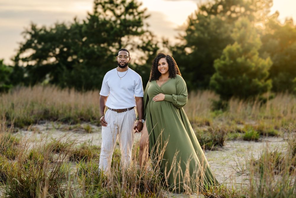 A couple in white and olive green outfits walk together in a sunlit meadow during golden hour with trees in background.