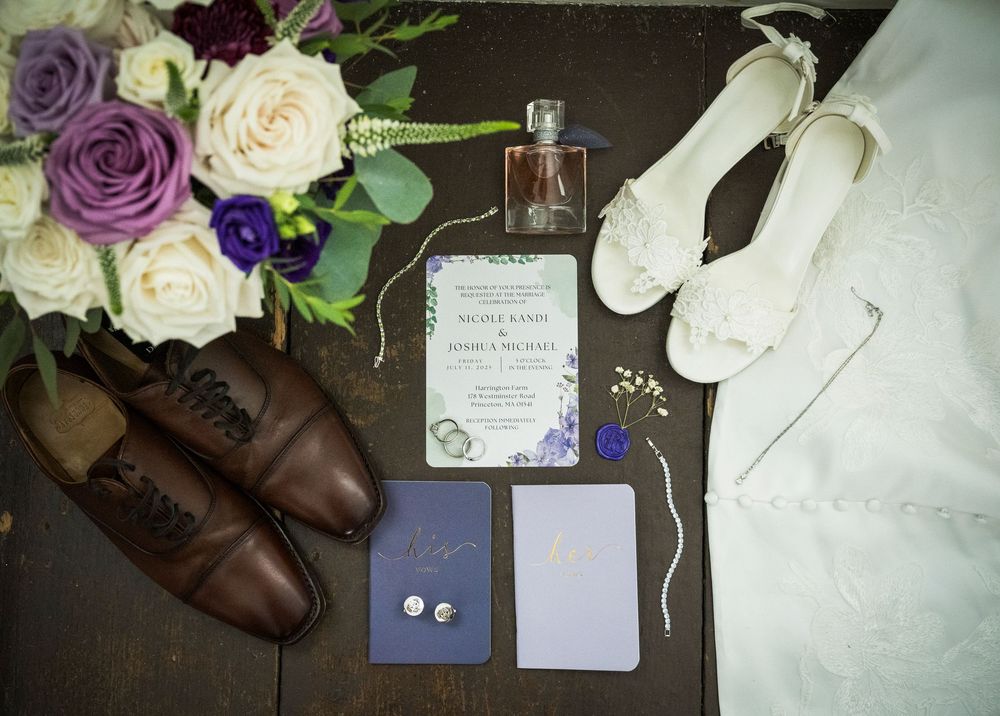 A wedding detail flatlay showing white and purple flowers, brown dress shoes, and blue invitation cards arranged artistically.