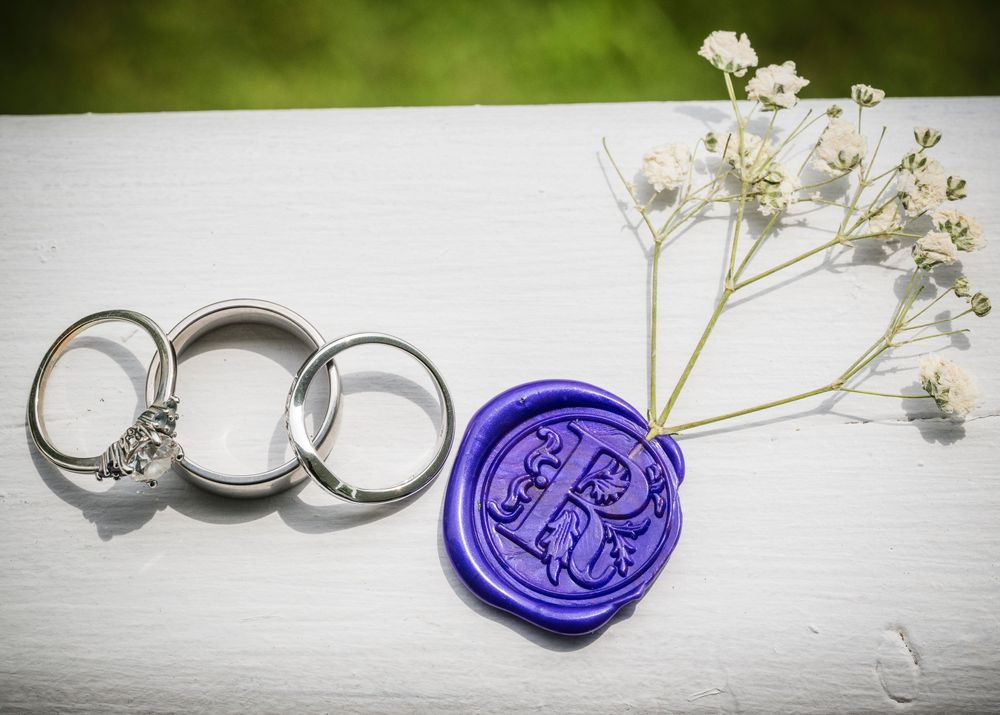 Wedding rings displayed next to a purple wax seal and delicate white baby's breath flowers on a white wooden surface.