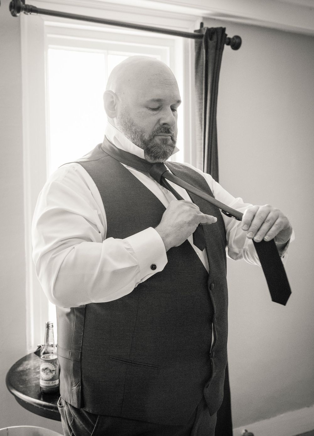 A person in formal attire adjusts their black necktie while wearing a dark vest and white dress shirt against a bright window.
