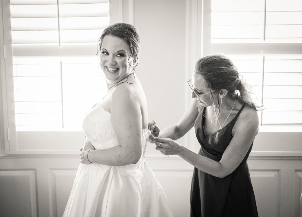 A wedding dress being fitted and adjusted in a bright sunlit room with window panels in the background.