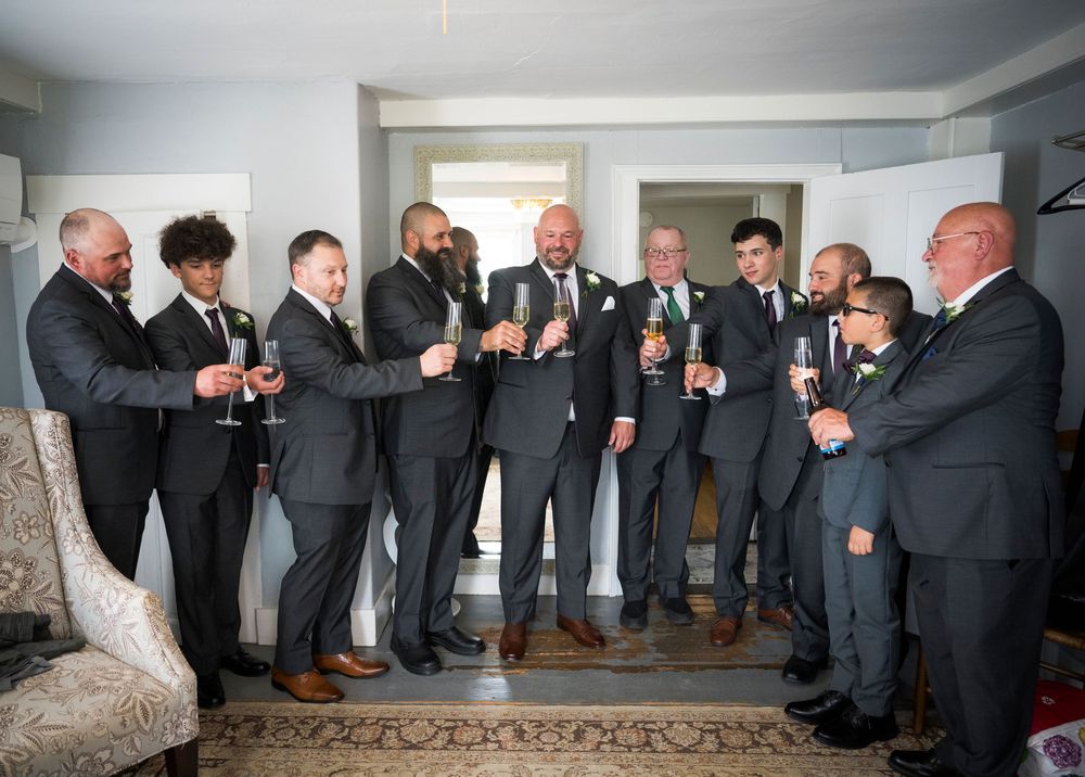 A group of groomsmen in dark suits share drinks and laughter while gathered together in a room.