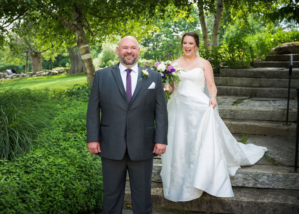A couple poses on garden steps for an outdoor wedding photo with lush greenery surrounding them during a summer ceremony.