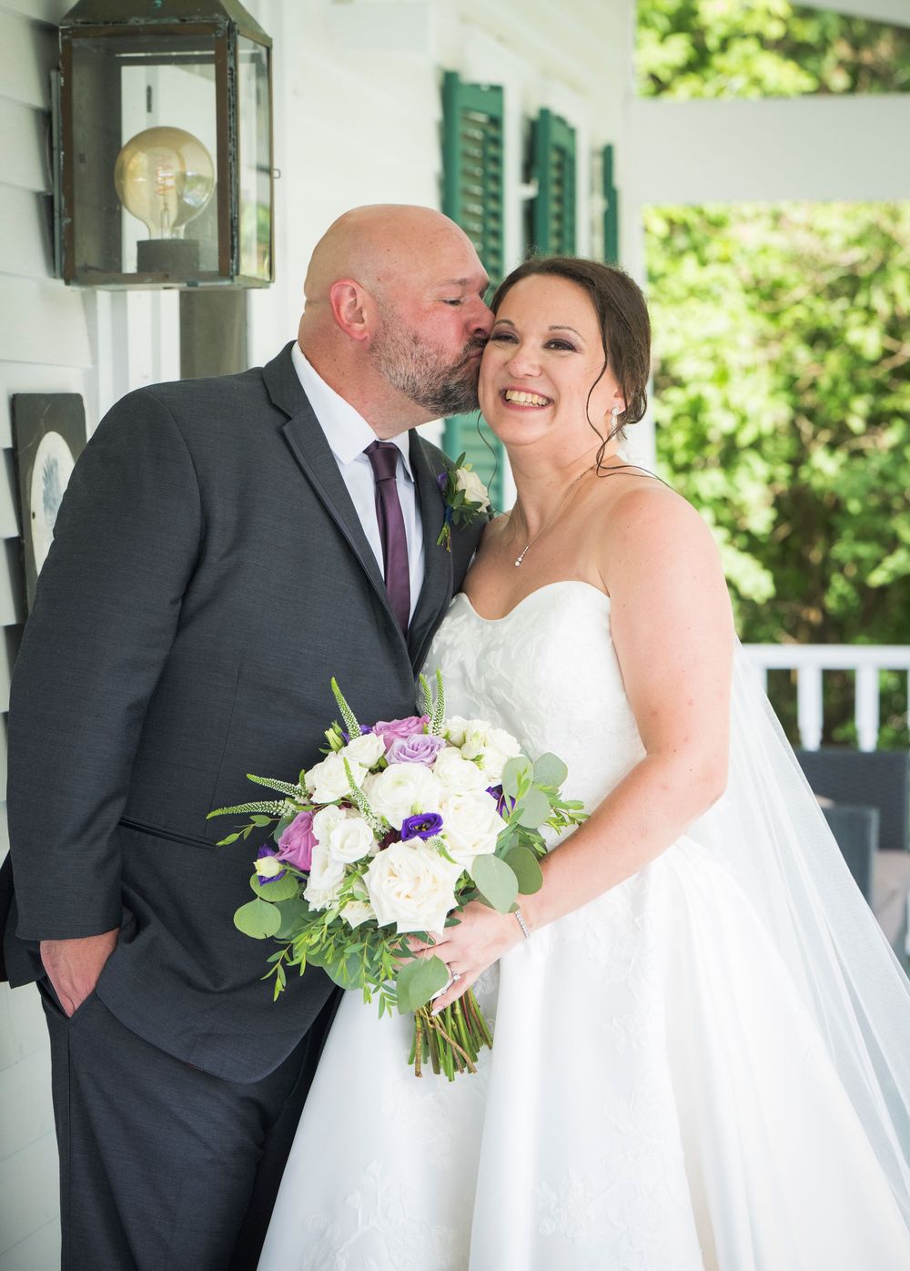 A joyful wedding couple embraces outdoors with a beautiful white and purple floral bouquet on a sunny day.