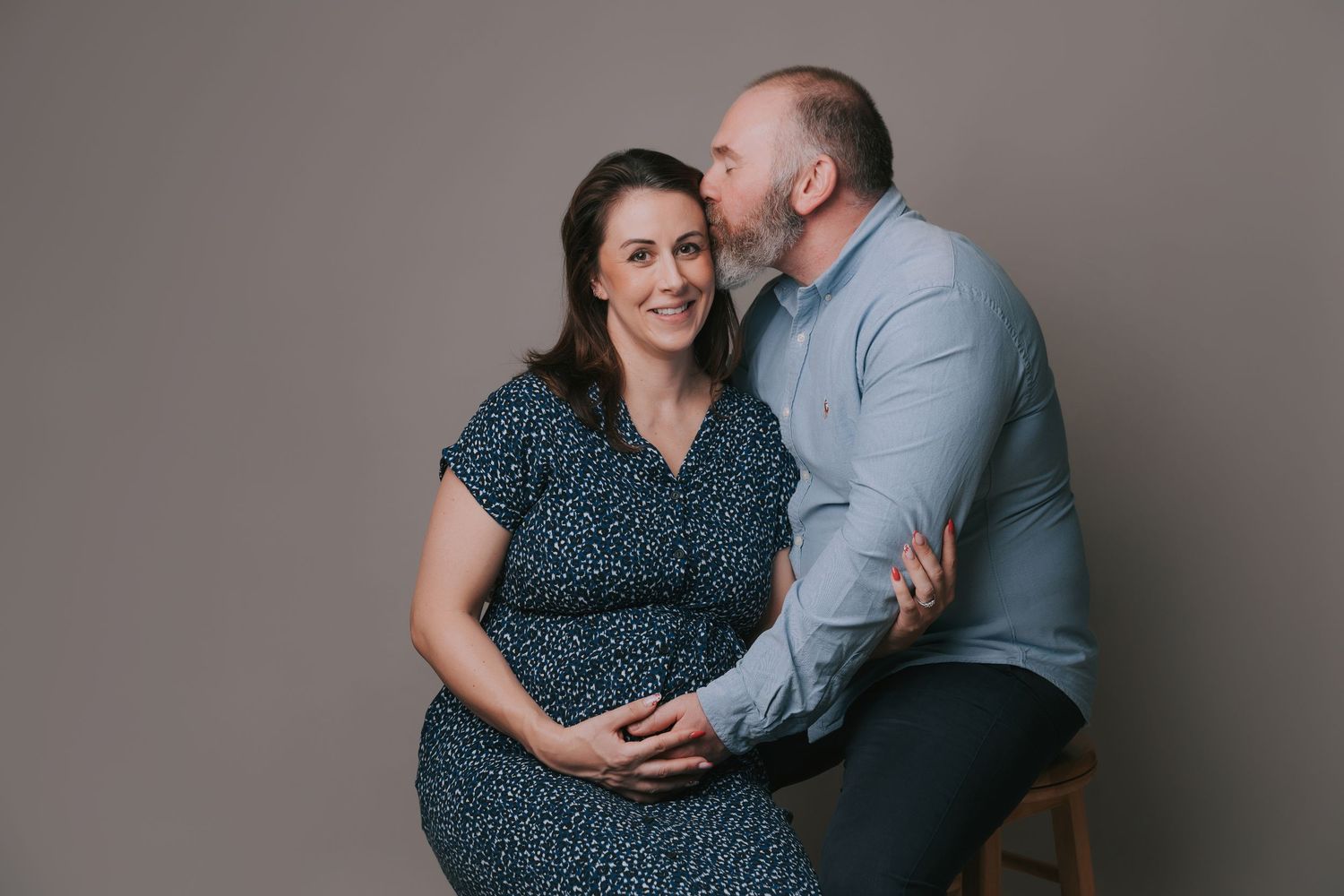 A man kisses a smiling pregnant woman on the forehead as they pose affectionately together for a maternity portrait.