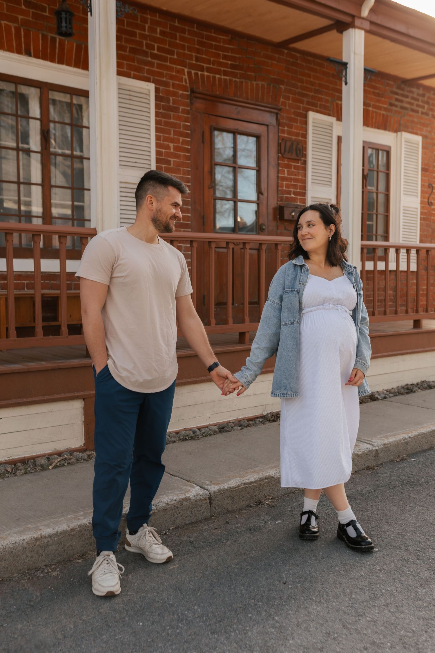 Expecting couple holding hands walking in front of red brick building at sunset.