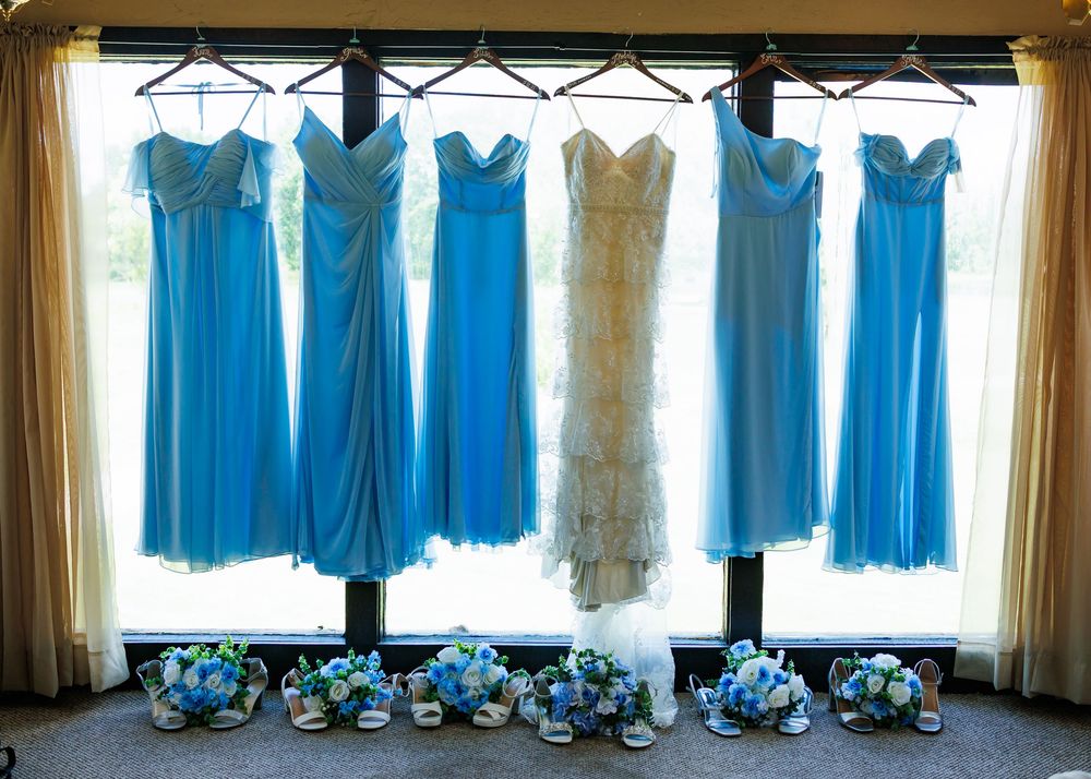 A row of light blue and cream bridesmaid dresses hanging on display against windows with sheer curtains.