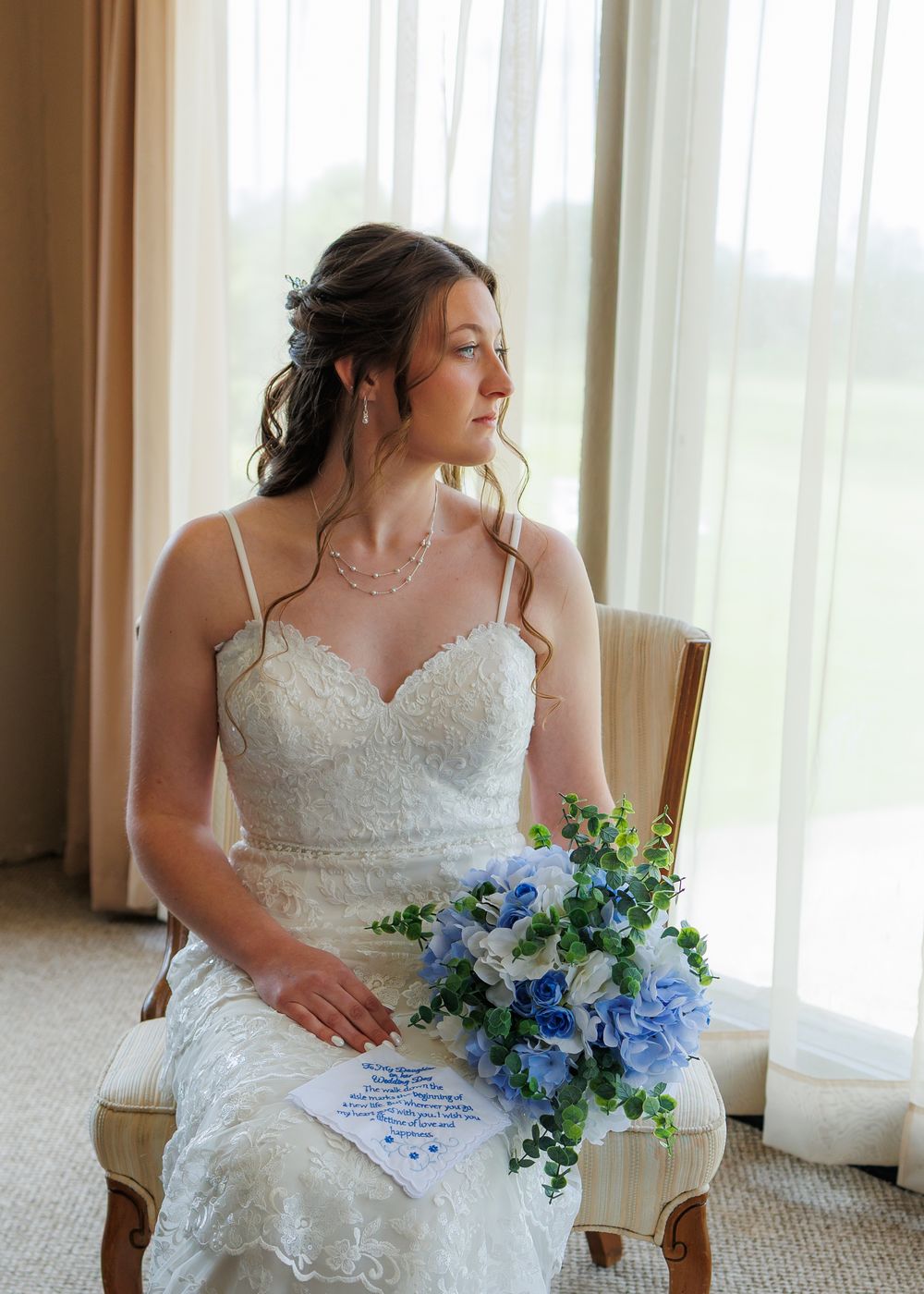 A bride in a white lace wedding dress sits by a window holding a blue and white floral bouquet.