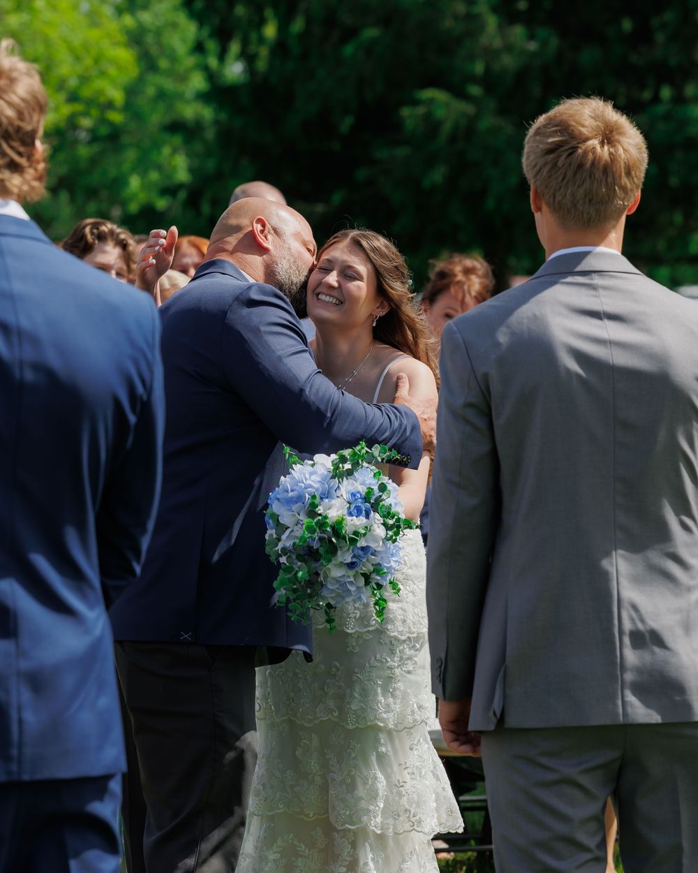 A bride in a white lace dress holds a blue and white bouquet while celebrating during an outdoor wedding ceremony.
