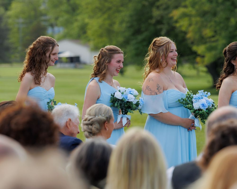 Bridesmaids in light blue dresses holding bouquets stand together during an outdoor wedding ceremony.