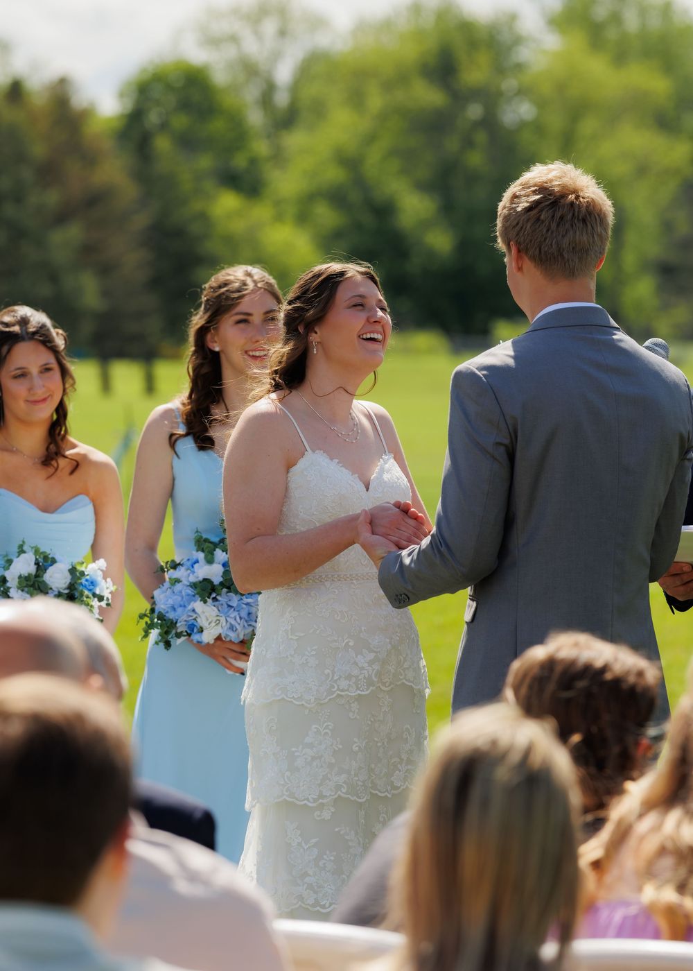 A joyful outdoor wedding ceremony with bridesmaids in light blue dresses holding blue bouquets on a sunny spring day.