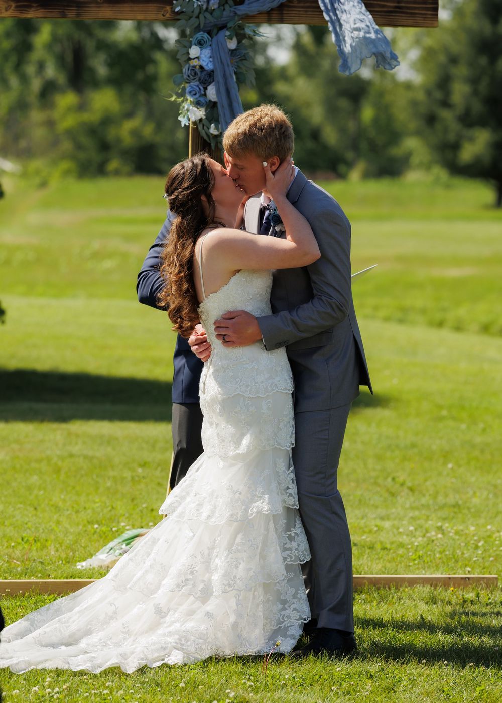 A couple shares their first kiss during an outdoor wedding ceremony on a sunny lawn beneath blue decorations.