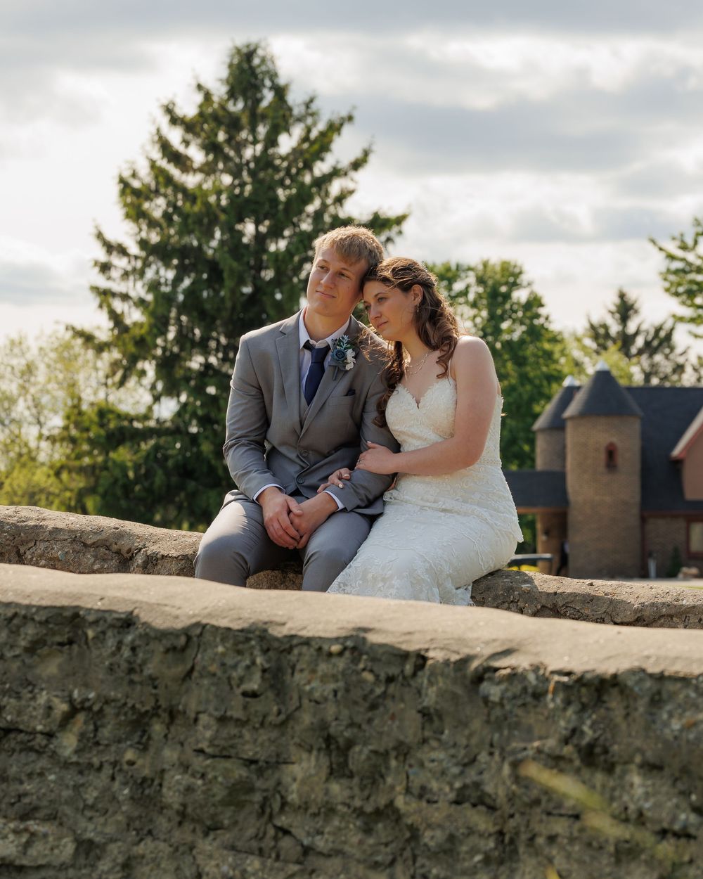 A romantic wedding couple sits together on an old stone wall with historic architecture and evergreen trees in the background.