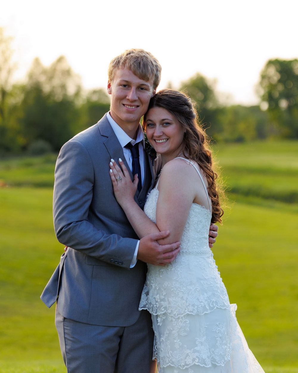 A couple in wedding attire embraces on a lush green golf course during sunset while showing off their rings.