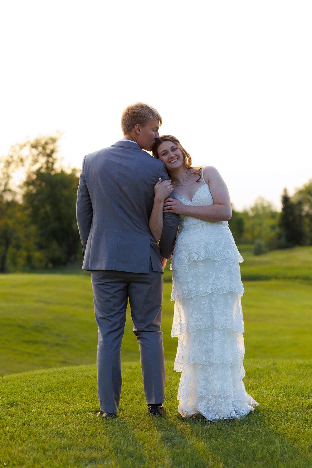 A romantic wedding couple embraces on a lush green lawn during golden hour sunset.