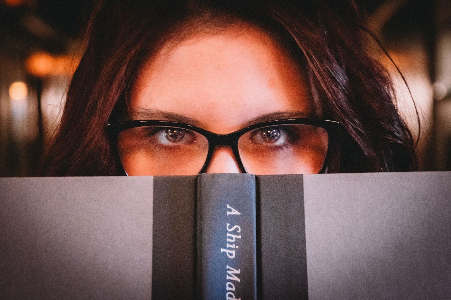 A close-up shot of eyes peering over a book in atmospheric lighting.