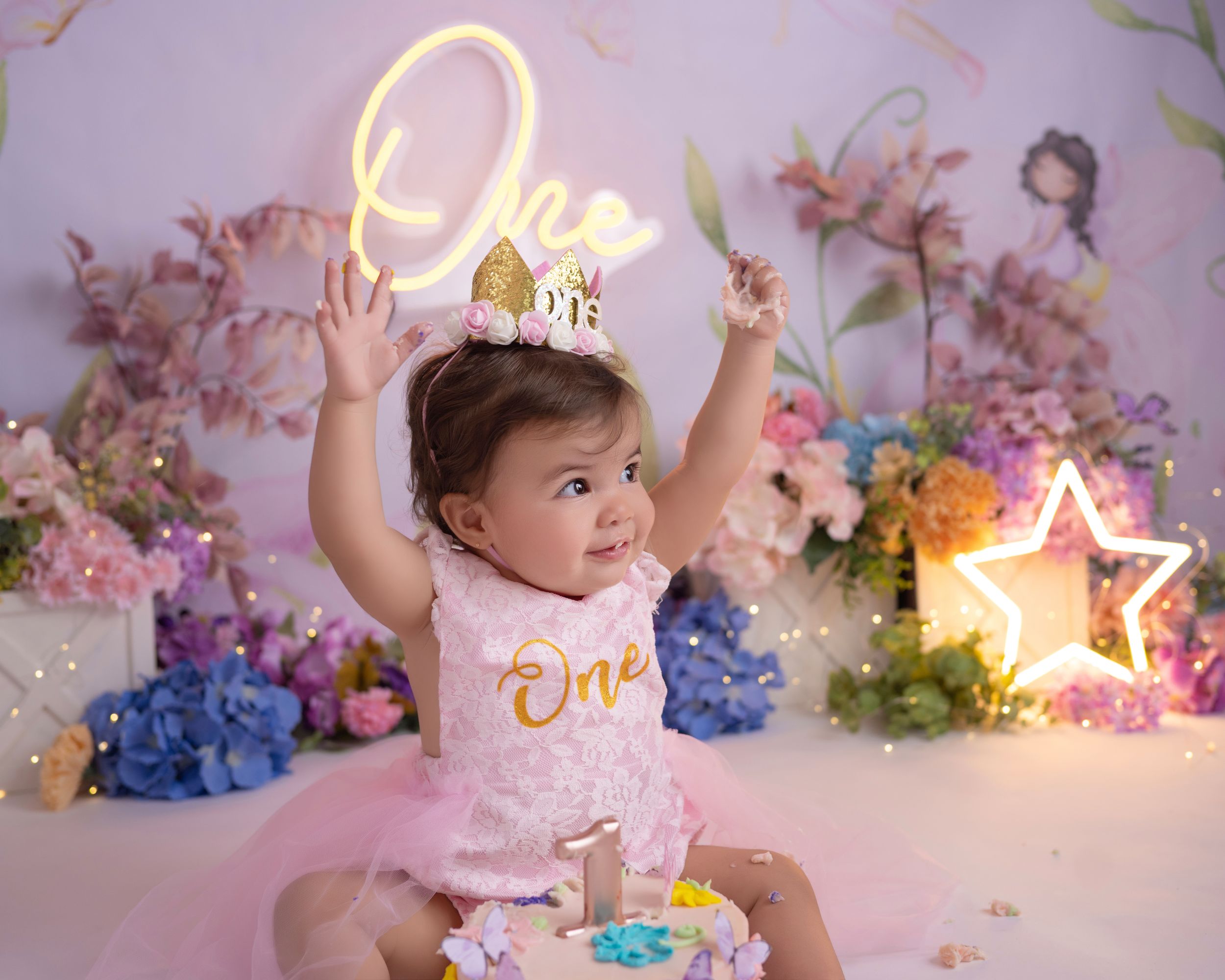 Floral birthday setup with neon 'One' sign and star light against pink backdrop.
