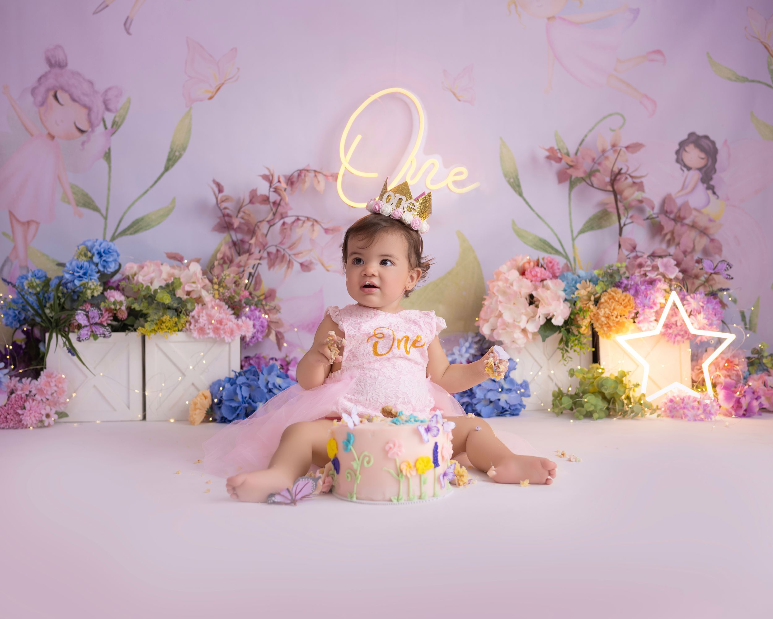 First birthday setup with floral arrangements and illuminated 'One' sign against pink backdrop.