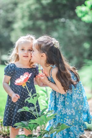 Two young children in patterned dresses share a secret while standing next to a blooming pink flower in a garden.