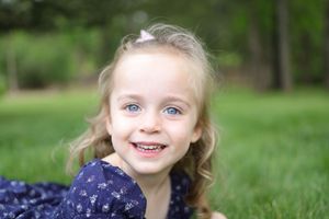 Young child wearing a navy polka dot dress smiles joyfully while playing outdoors in a grassy field.