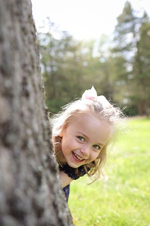 A young child peers playfully from behind a tree trunk in a sunny outdoor setting with green grass and trees.