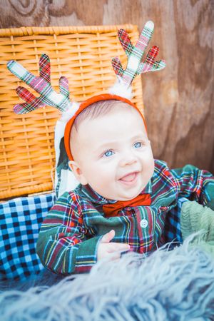 Baby wearing festive plaid outfit with reindeer antler headband sits by wicker basket on blue checkered blanket.