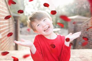 Child in red sweater plays joyfully with falling red rose petals against a soft blurred background.