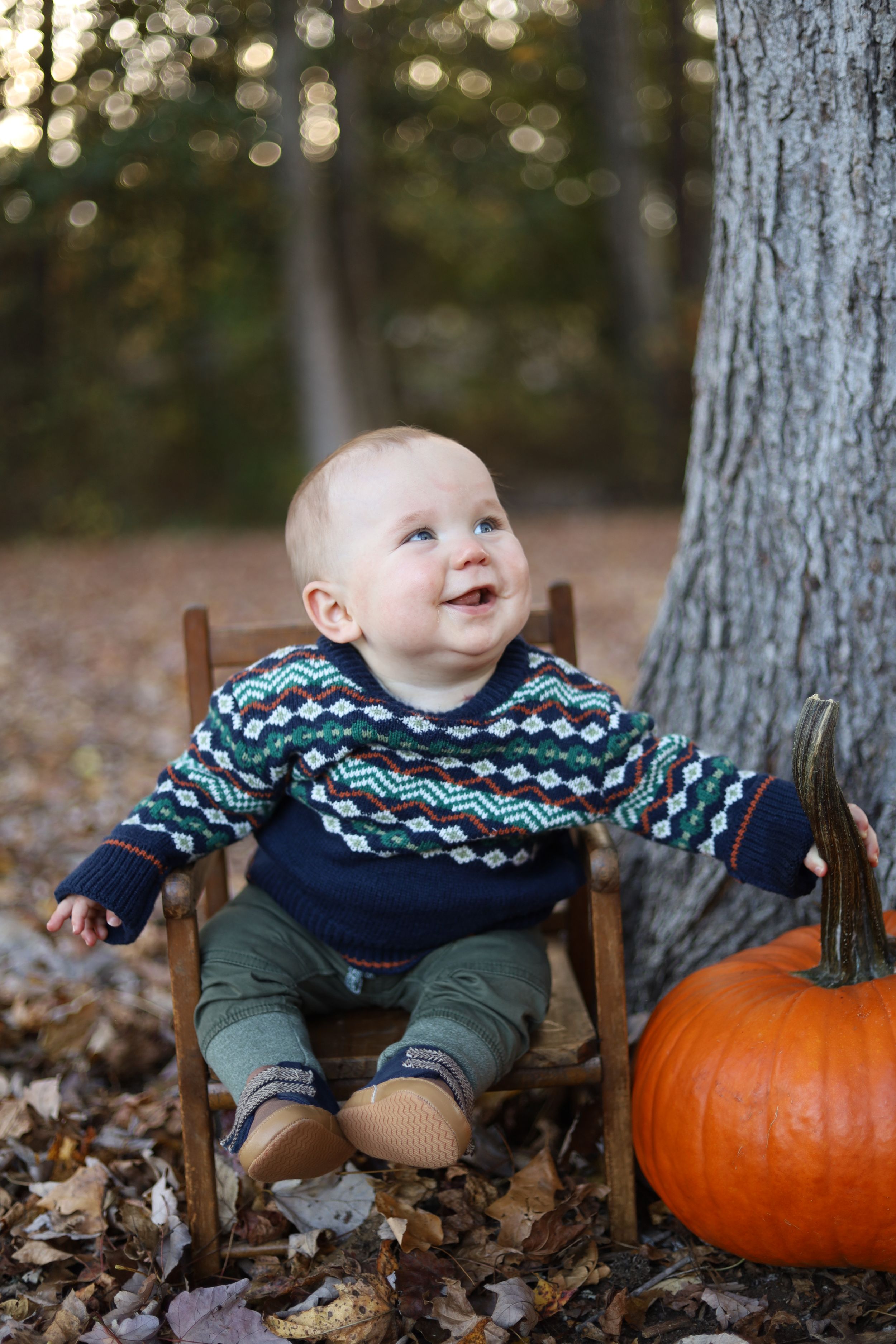 Baby in fall sweater sits in wooden chair next to pumpkin surrounded by autumn leaves on forest floor.