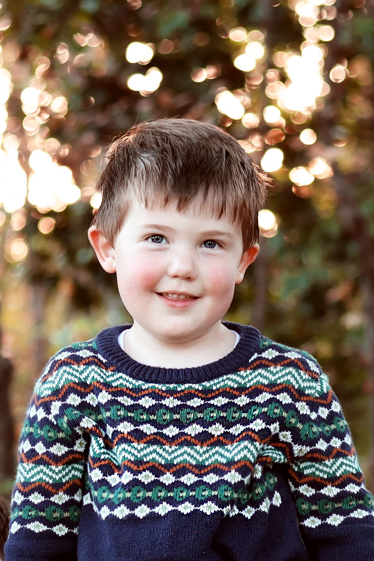 A young child wearing a navy fair isle sweater smiles outdoors with bokeh lights in the background.