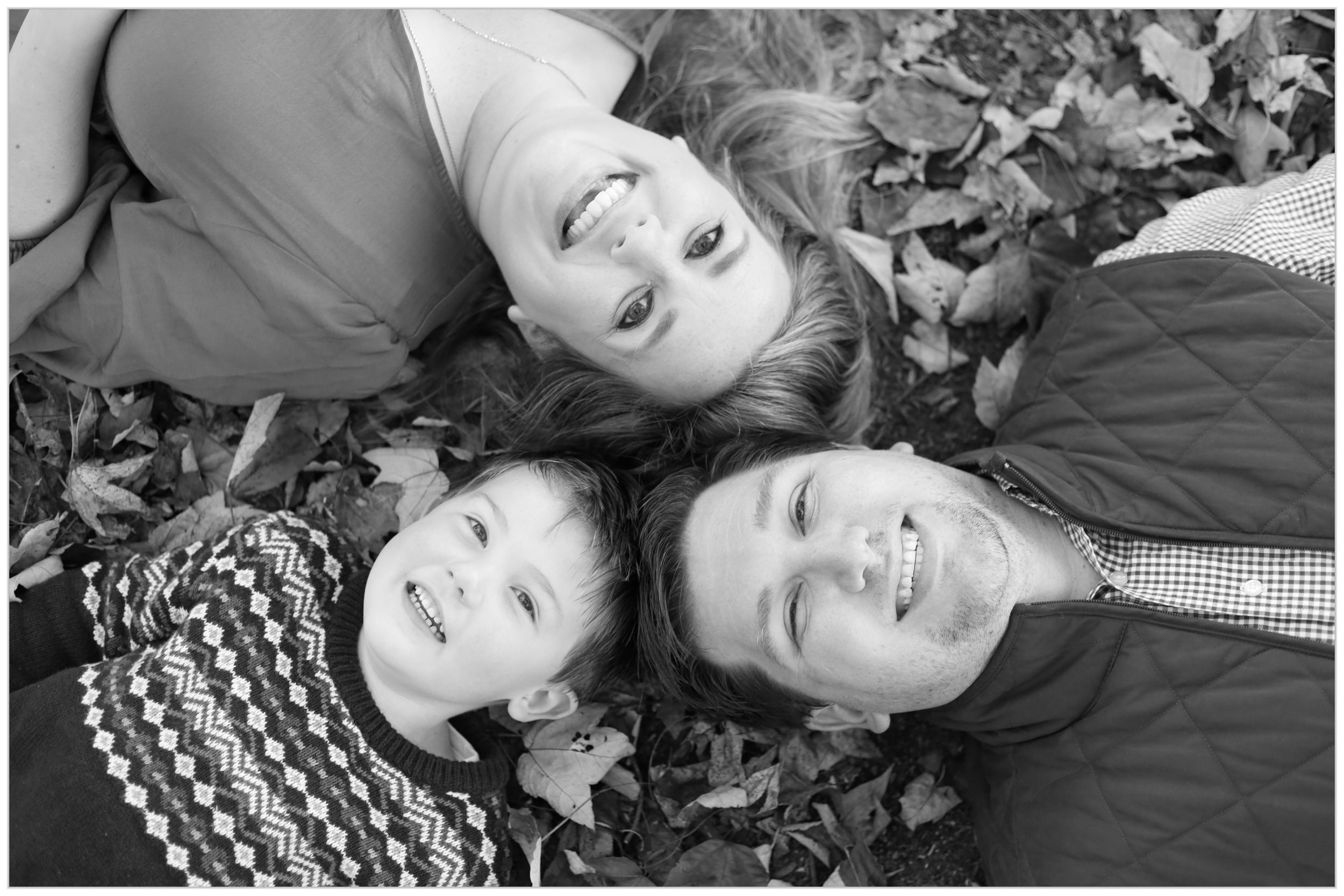 Black and white photo of three people lying on autumn leaves with their heads together while smiling upward.