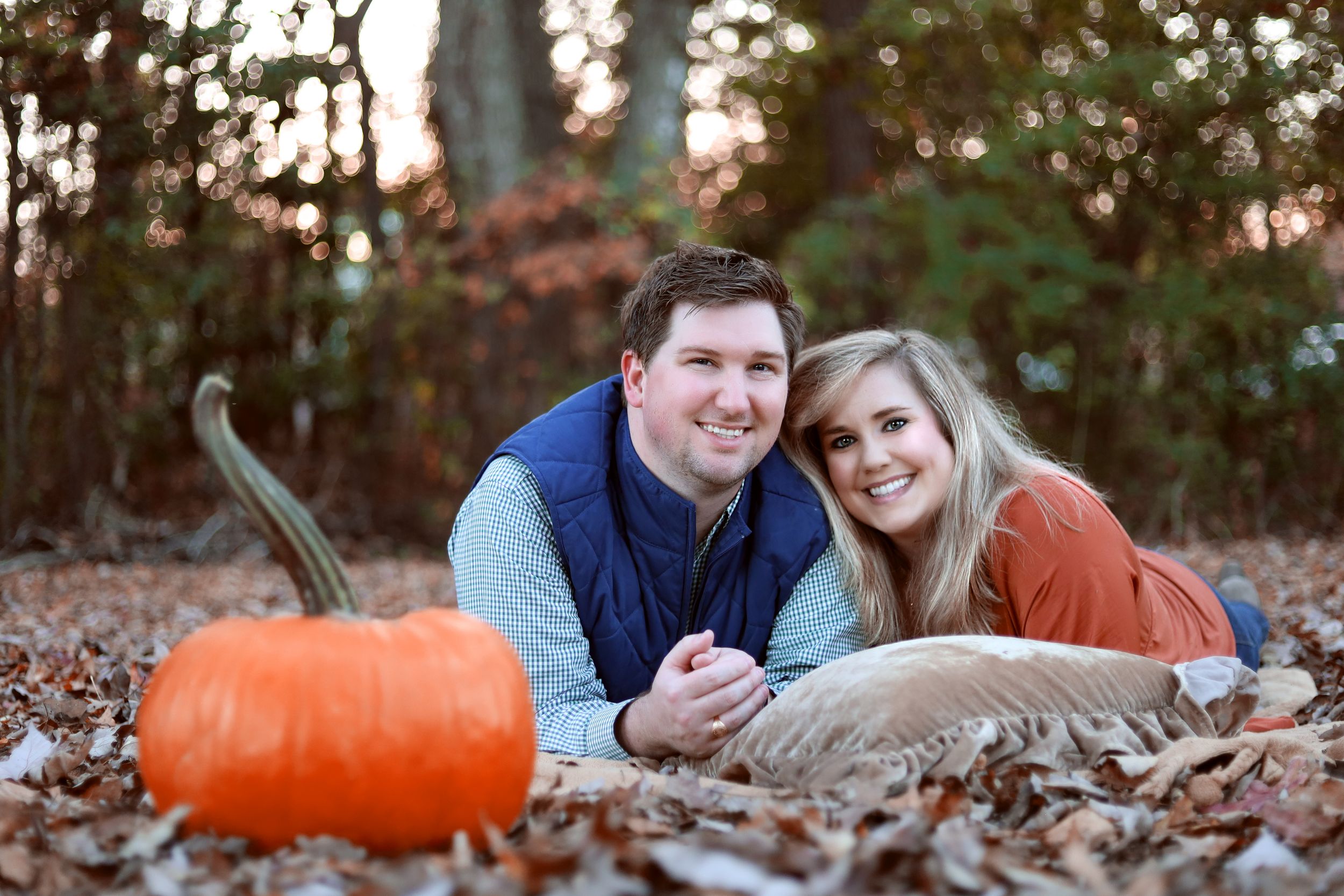 A couple lies in autumn leaves next to a large orange pumpkin during an outdoor fall photo session.