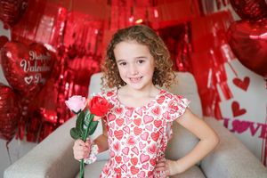 Heart-themed Valentine's Day photo setup with red balloons and decorations featuring a heart-patterned dress and red rose.