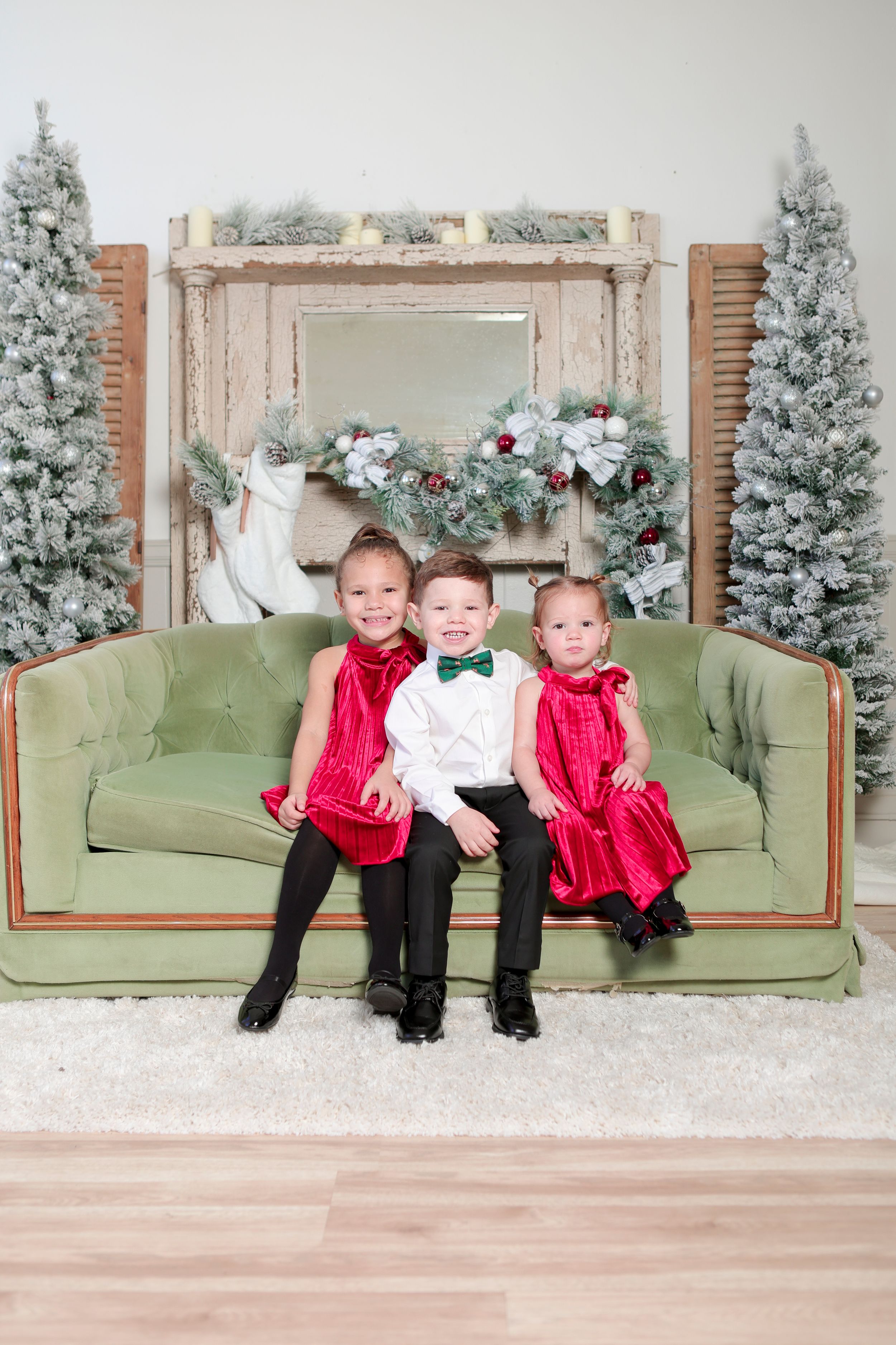 A festive holiday scene with three children sitting on a green velvet couch surrounded by decorated Christmas trees and garland.